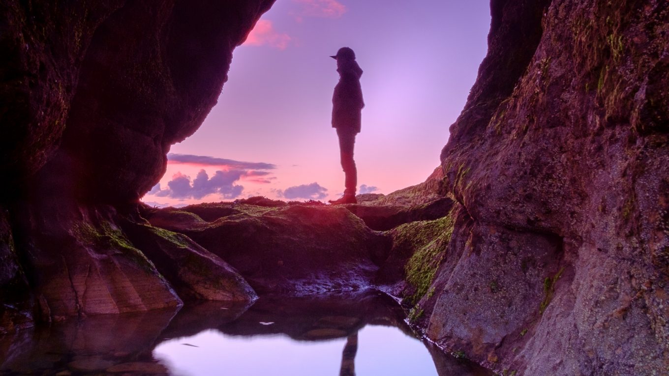 person standing in cave entrance with reflection in pool of water