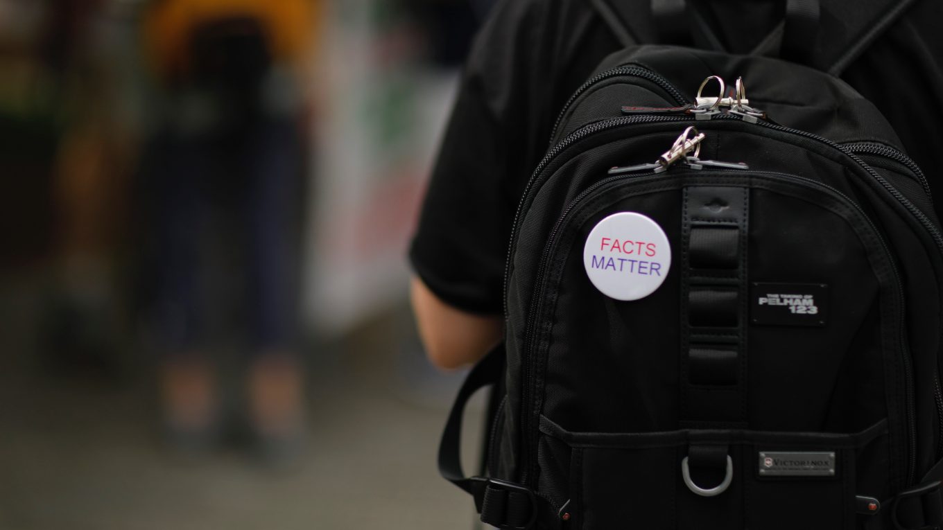 A white“Facts matter” pin on a black backpack on a person's back